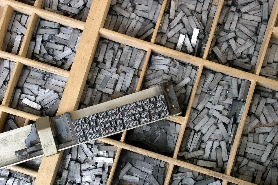 A wooden type case filled with small metal letters, a composing stick of set type lying across it